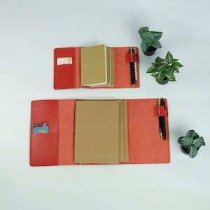 Puede incluir: Dos cubiertas de diario de cuero rojo abiertas con cuadernos de papel marrón en el interior. Cada cubierta tiene un portalápices con un bolígrafo negro y un bolsillo con una tarjeta. Pequeñas plantas en macetas están en el fondo.