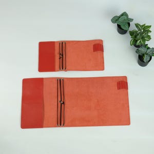 May include: Two open red leather journal covers with black elastic closures and a small pocket. The journals are displayed on a white surface. The covers are open, revealing the interior. The journals are in different sizes.