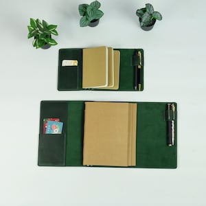 Puede incluir: Dos cubiertas de diario de cuero verde abiertas con cuadernos de papel marrón en el interior. Cada cubierta tiene un portalápices con un bolígrafo negro y un bolsillo para tarjetas. La cubierta superior está cerrada y la cubierta inferior está abierta. Pequeñas plantas en macetas en el fondo.