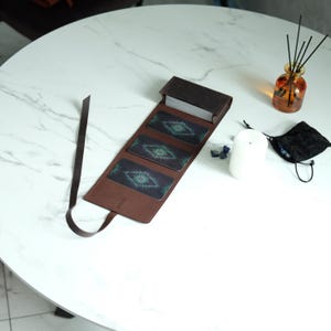 Peut inclure: Un étui à cartes de tarot en cuir marron avec une longue lanière, contenant un jeu de cartes et présentant trois cartes face visible. Chaque carte présente un motif graphique vert foncé et bleu sarcelle d'un œil stylisé avec des rayons.