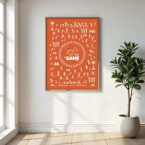 May include: An orange framed print with white illustrations of various sports figures. The text "Spirit of the Game" is in the center. The print is displayed on a white wall next to a potted plant and a window.