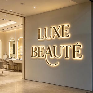 May include: A storefront entrance featuring the text LUXE BEAUTÉ in large, gold, backlit dimensional letters mounted on a smooth, off-white wall. Through the glass partition, a bright interior space with light-colored chairs and shelves is visible.