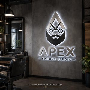 Może przedstawiać: Podświetlany szyld dla zakładu fryzjerskiego z napisem "APEX" w srebrnych literach. Znak zawiera stylizowaną ikonę brody i nożyczek. Poniżej znajduje się napis "BARBER STUDIO". Znak jest zamontowany na szarej ścianie.
