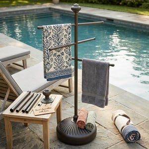 May include: A bronze-colored towel rack with multiple horizontal bars, holding several towels of various colors and patterns. The rack stands on a round base near a swimming pool. A small wooden table with the text "Hand-Assembled with Care" is next to the rack.