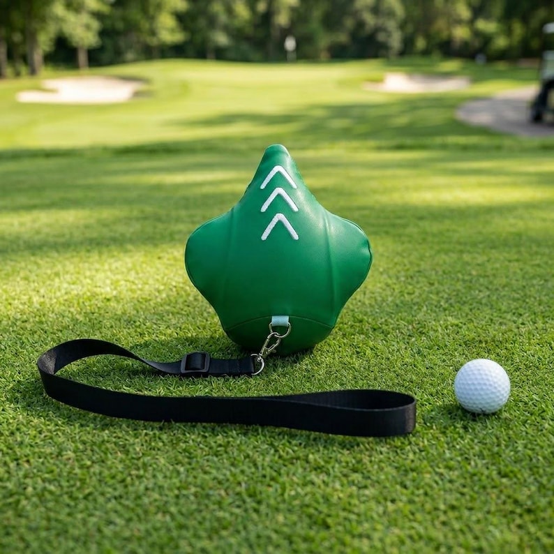May include: A green golf swing training aid with white arrow markings and a black strap, next to a white golf ball, all on a green golf course.