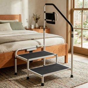 May include: A white metal two-step stool with black non-slip treads and a tall, integrated support handle featuring a black foam grip and a small black storage pouch. The frame is adjustable in height and designed for bedside assistance.