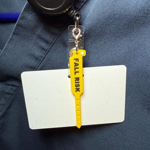 May include: A yellow plastic tag with the black text FALL RISK printed vertically, attached to a white rectangular identification card holder. The tag is connected to a black retractable badge reel clipped onto a dark blue fabric garment.