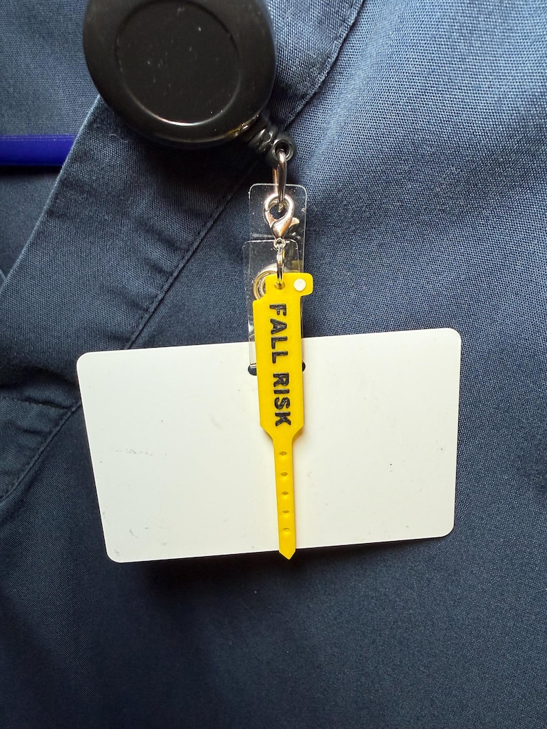 May include: A yellow plastic tag with the black text FALL RISK printed vertically, attached to a white rectangular identification card holder. The tag is connected to a black retractable badge reel clipped onto a dark blue fabric garment.