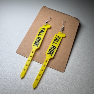 May include: A pair of yellow earrings with the text "FALL RISK" in black. The earrings are shaped like medical wristbands and have small holes along the length. They are displayed on a brown card.