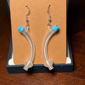 Puede incluir: Un par de pendientes claros y curvos con detalles azules en la parte superior y detalles en forma de corazón en la parte inferior. Los pendientes se exhiben en una tarjeta marrón dentro de una caja azul, lo que sugiere que son una joya hecha a mano.