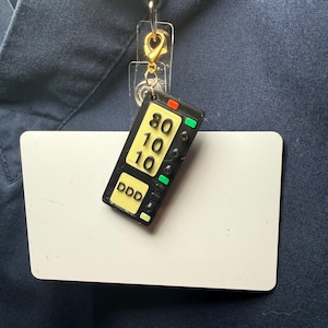 May include: A black and yellow remote control badge reel charm with the numbers 80, 10, and 10, and the letters DDD. The charm is attached to a clear plastic badge holder with a gold-colored clasp and a black retractable reel.