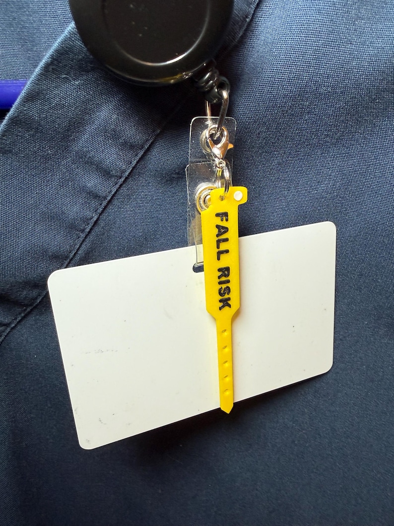 May include: A yellow plastic tag with the text FALL RISK printed in black, attached to a clear plastic badge holder and a black retractable reel. The badge holder is blank and white, and the entire assembly is clipped to a dark blue fabric garment.