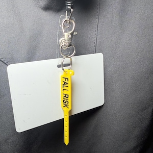 May include: A yellow "FALL RISK" tag attached to a white ID badge. The tag is connected to a retractable badge reel with a metal clip. The background is a gray fabric.