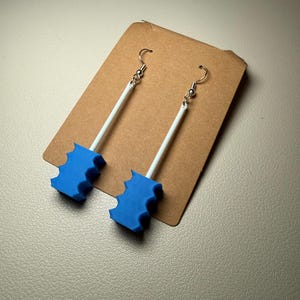 May include: A pair of handmade earrings featuring blue, bat-shaped charms. The charms are attached to white rods and silver-colored ear wires. The earrings are displayed on a brown card.