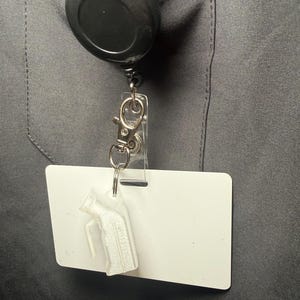 Könnte beinhalten: Eine schwarze, einziehbare Ausweisrolle mit einem transparenten Ausweishalter und einer weißen Ausweiskarte. Ein kleines, weißes, 3D-gedrucktes Objekt hängt am Ausweishalter. Die Rolle ist an einem dunkelgrauen Kleidungsstück befestigt.