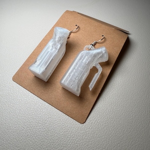 May include: A pair of white, 3D-printed earrings shaped like small spray bottles. The earrings are hanging on silver-colored hooks and are displayed on a brown cardboard backing.
