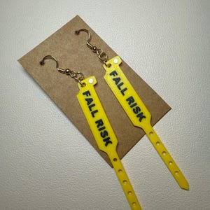 Pode incluir: Um par de brincos pendentes amarelos em forma de pulseiras de identifica&ccedil;&atilde;o hospitalar. Cada brinco apresenta o texto FALL RISK impresso em letras mai&uacute;sculas pretas em negrito, preso a ganchos de metal dourado para orelhas furadas.
