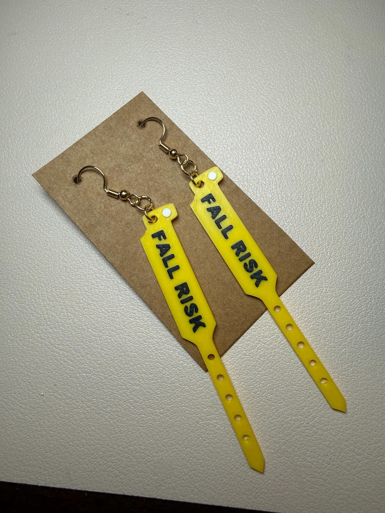 Pode incluir: Um par de brincos pendentes amarelos em forma de pulseiras de identifica&ccedil;&atilde;o hospitalar. Cada brinco apresenta o texto FALL RISK impresso em letras mai&uacute;sculas pretas em negrito, preso a ganchos de metal dourado para orelhas furadas.