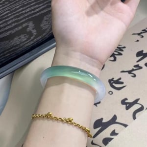 Può includere: Un bracciale a cerchio in giada nei toni del verde e del bianco è indossato su un polso, abbinato a un braccialetto a catena d'oro. Il bracciale in giada ha una superficie liscia e lucida. Il braccialetto d'oro presenta piccoli ciondoli decorativi.