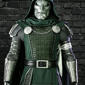 May include: A full-body costume featuring a metallic silver mask and hood, a dark green tunic with a textured square pattern, silver shoulder pauldrons, chain-linked chest clasps, black gauntlets, and a black belt with a silver buckle.