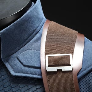 May include: Close-up of a blue and brown costume. The blue fabric has a textured appearance with a padded shoulder detail. A brown strap with a silver buckle crosses the shoulder. The costume is displayed on a black mannequin.