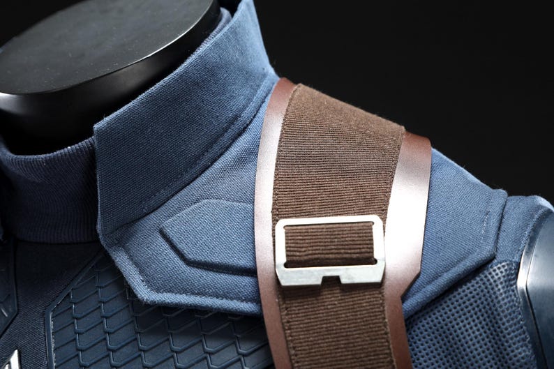 May include: Close-up of a blue and brown costume. The blue fabric has a textured appearance with a padded shoulder detail. A brown strap with a silver buckle crosses the shoulder. The costume is displayed on a black mannequin.