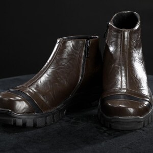 May include: A pair of brown leather boots with a zip fastening. The boots have a rugged, black sole and a black accent stripe across the toe. The boots are displayed on a dark surface.