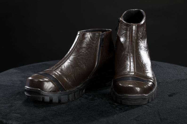May include: A pair of brown leather boots with a zip fastening. The boots have a rugged, black sole and a black accent stripe across the toe. The boots are displayed on a dark surface.