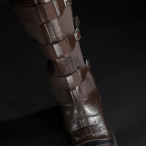 May include: Brown leather boot with multiple straps and buckles. The boot has a dark brown sole and a textured surface. The leg is covered with a brown fabric. The boot is worn with blue jeans.