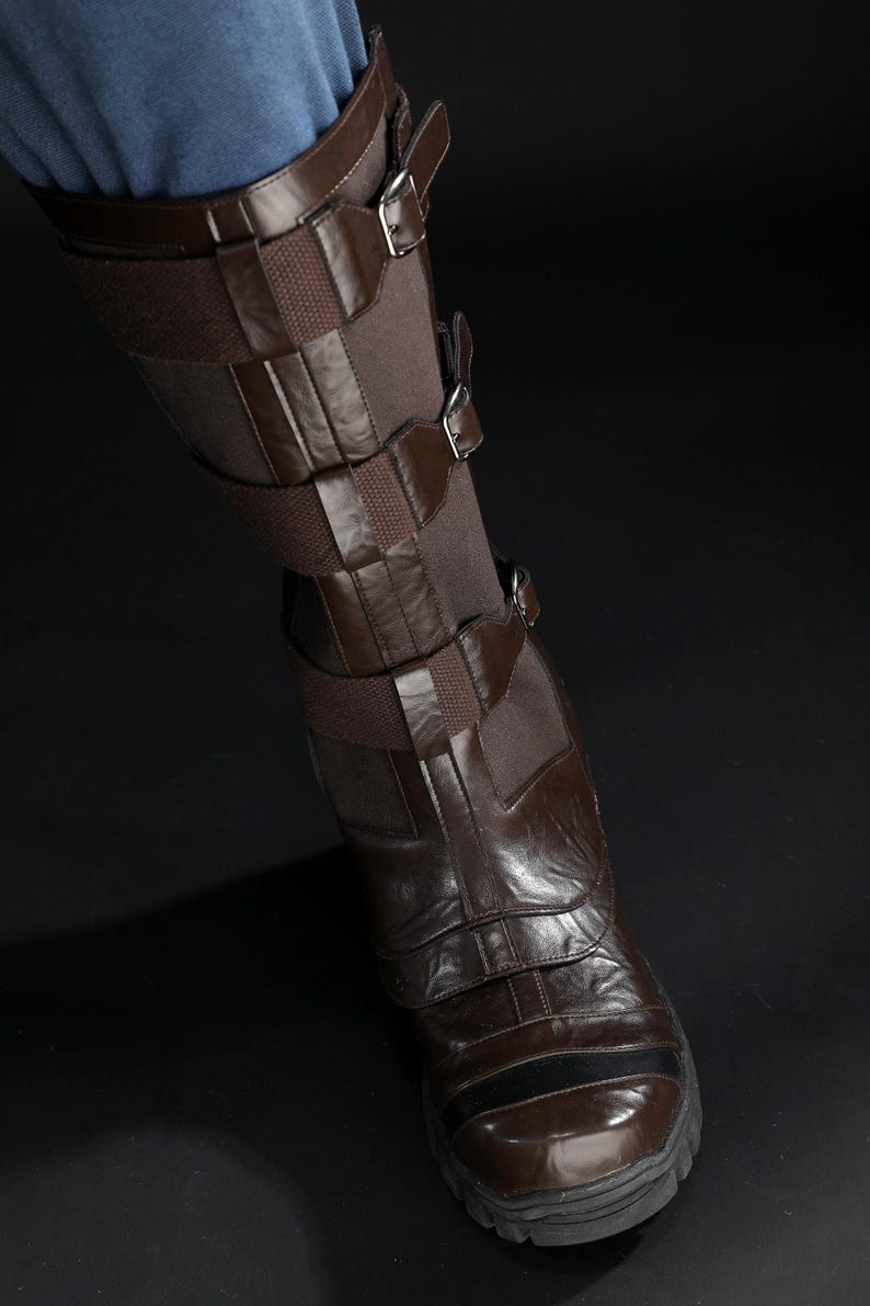 May include: Brown leather boot with multiple straps and buckles. The boot has a dark brown sole and a textured surface. The leg is covered with a brown fabric. The boot is worn with blue jeans.