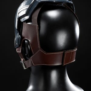 May include: A dark blue and silver helmet with a brown leather and fabric harness. The helmet is displayed on a black mannequin head. The harness has white stitching and a metal buckle.
