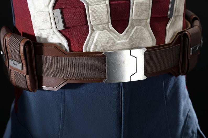 May include: A close-up of a brown utility belt with a silver buckle. The belt has two pouches and is worn over a red, white, and blue costume. The buckle is rectangular with a geometric design.