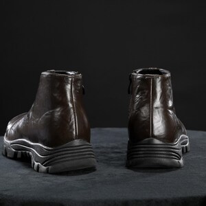 May include: Two brown leather ankle boots with a glossy finish are shown from the back. The boots have a zip at the back and a thick, ridged sole. They are set against a dark background.