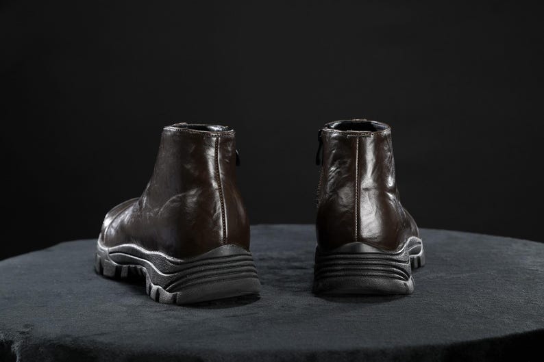 May include: Two brown leather ankle boots with a glossy finish are shown from the back. The boots have a zip at the back and a thick, ridged sole. They are set against a dark background.