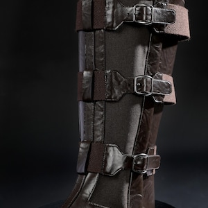 May include: Brown leather leg armour with multiple straps and buckles. The armour has a dark brown colour with lighter brown accents. The design includes several horizontal straps secured with metal buckles, and a dark fabric panel.