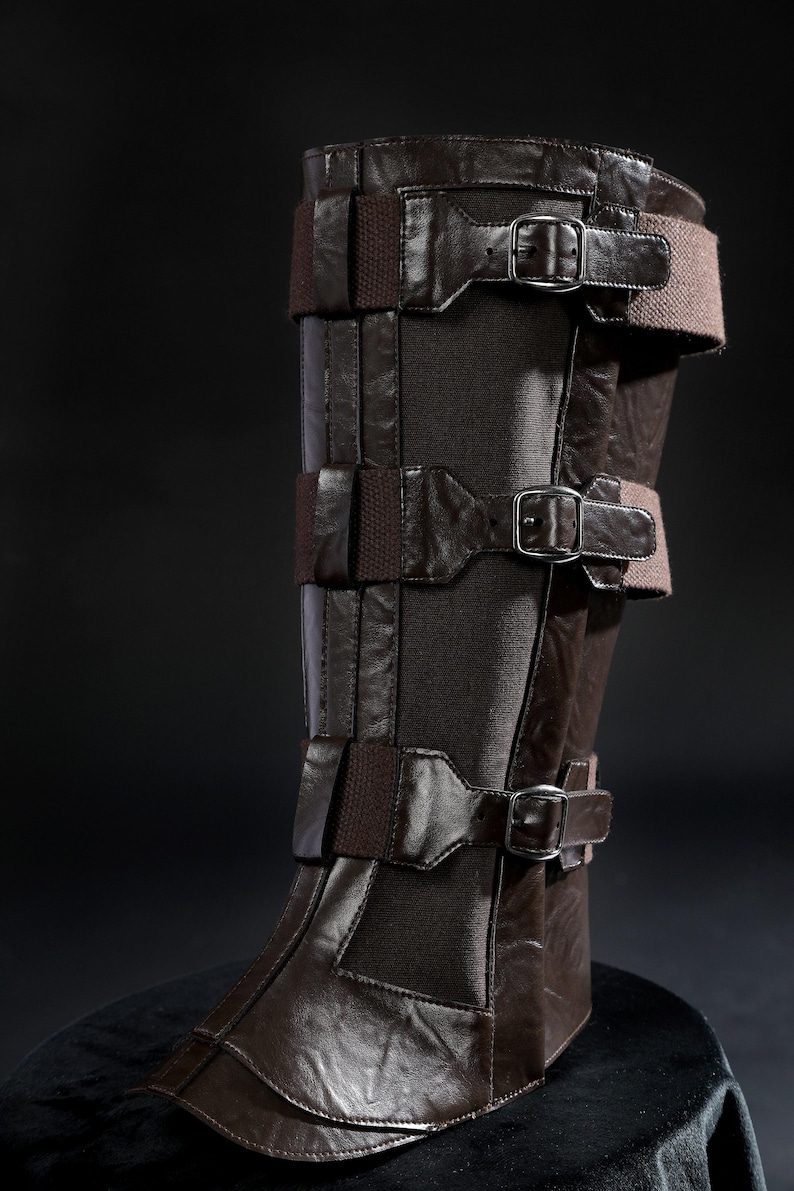 May include: Brown leather leg armour with multiple straps and buckles. The armour has a dark brown colour with lighter brown accents. The design includes several horizontal straps secured with metal buckles, and a dark fabric panel.