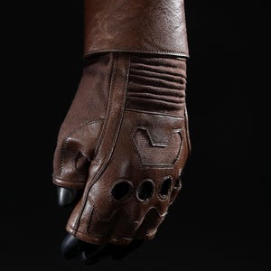 May include: Brown leather glove with fingerless design and ventilation holes. The glove features a padded wrist cuff and a textured fabric on the back of the hand. The fingers have black tips. The glove is worn over a blue sleeve.