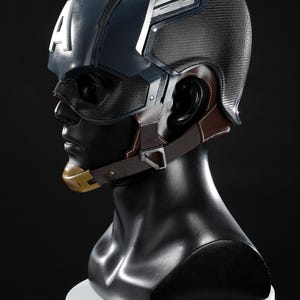 May include: A Captain America helmet replica, displayed on a black mannequin head. The helmet is navy blue with white accents and a large white "A" on the side. It features a mesh face covering, brown ear and chin straps, and a gold chin piece.
