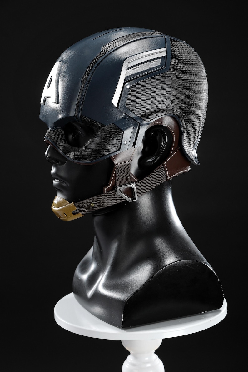 May include: A Captain America helmet replica, displayed on a black mannequin head. The helmet is navy blue with white accents and a large white "A" on the side. It features a mesh face covering, brown ear and chin straps, and a gold chin piece.