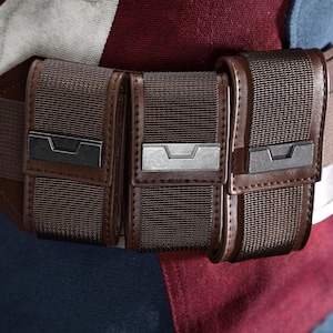 May include: A close-up of a brown utility belt with three pouches. The belt is made of a dark brown material with a matching buckle. The pouches have a textured, woven appearance and are secured with metal clasps.