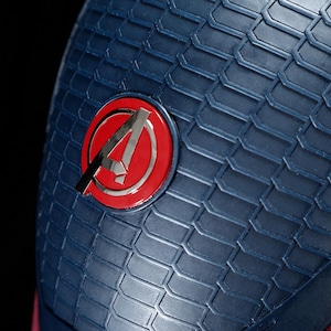 May include: Close-up of a blue textured surface with a red and silver Avengers logo. The logo is a stylized letter "A" inside a red circle. The surface has a geometric pattern.