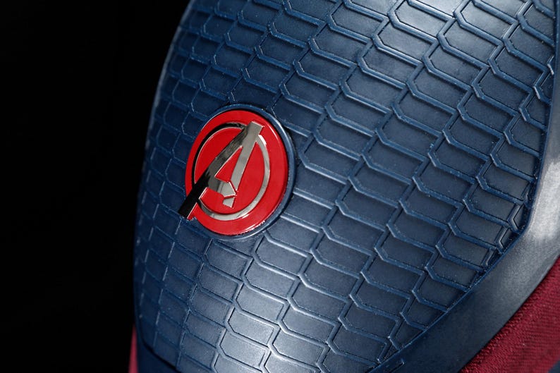 May include: Close-up of a blue textured surface with a red and silver Avengers logo. The logo is a stylized letter "A" inside a red circle. The surface has a geometric pattern.