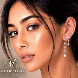 May include: A set of gold-toned earrings featuring small light blue floral-shaped accents and clear stones. The jewellery includes a single stud, a curved bar with a floral charm, and a long drop earring with multiple floral accents and a pear-shaped stone.