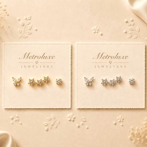 May include: Two square display cards labelled Metroluxe Jewellery, each holding three stud earrings. The left card features gold-tone butterfly, triple-star, and round solitaire earrings. The right card displays matching silver-tone versions of the same designs.