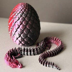 May include: A 3D-printed dragon egg and articulated dragon toy. The egg is red and black with a scaled texture. The dragon is also red and black, with a segmented body and a dragon head.