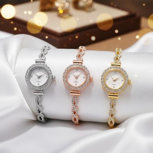 Puede incluir: Tres relojes de cuarzo con biseles con incrustaciones de cristal y correas de pulsera con eslabones. Las esferas son blancas. Un reloj es plateado, otro es de oro rosa y otro es dorado. Se presentan sobre una tela blanca satinada.