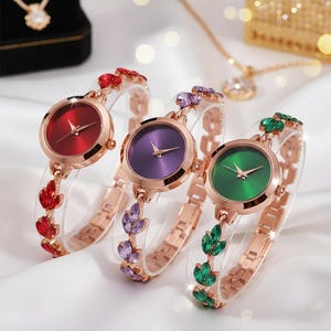 Puede incluir: Tres relojes de oro rosa con esferas coloridas y correas decorativas. Las esferas son rojas, moradas y verdes. Las correas presentan plástico transparente y detalles de piedras preciosas a juego. Los relojes se exhiben sobre una superficie blanca.