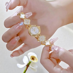 Puede incluir: Un reloj de pulsera dorado con esfera blanca y agujas finas. La correa presenta eslabones en forma de trébol con cristales transparentes y detalles de nácar. El reloj es sostenido por una persona.