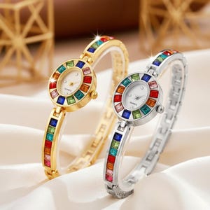 Puede incluir: Dos relojes de cuarzo con detalles de piedras preciosas de colores. Uno es dorado, el otro plateado. Ambos tienen esferas blancas y pulseras de piedras preciosas a juego. Los relojes se exhiben sobre una superficie sedosa blanca.