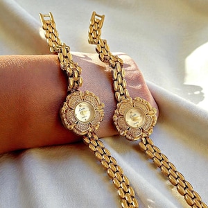 Puede incluir: Dos relojes de cuarzo dorados con esferas en forma de flor y correas de eslabones. Las esferas son blancas con agujas doradas y la marca "Luter". Las correas están hechas de pequeños eslabones dorados.